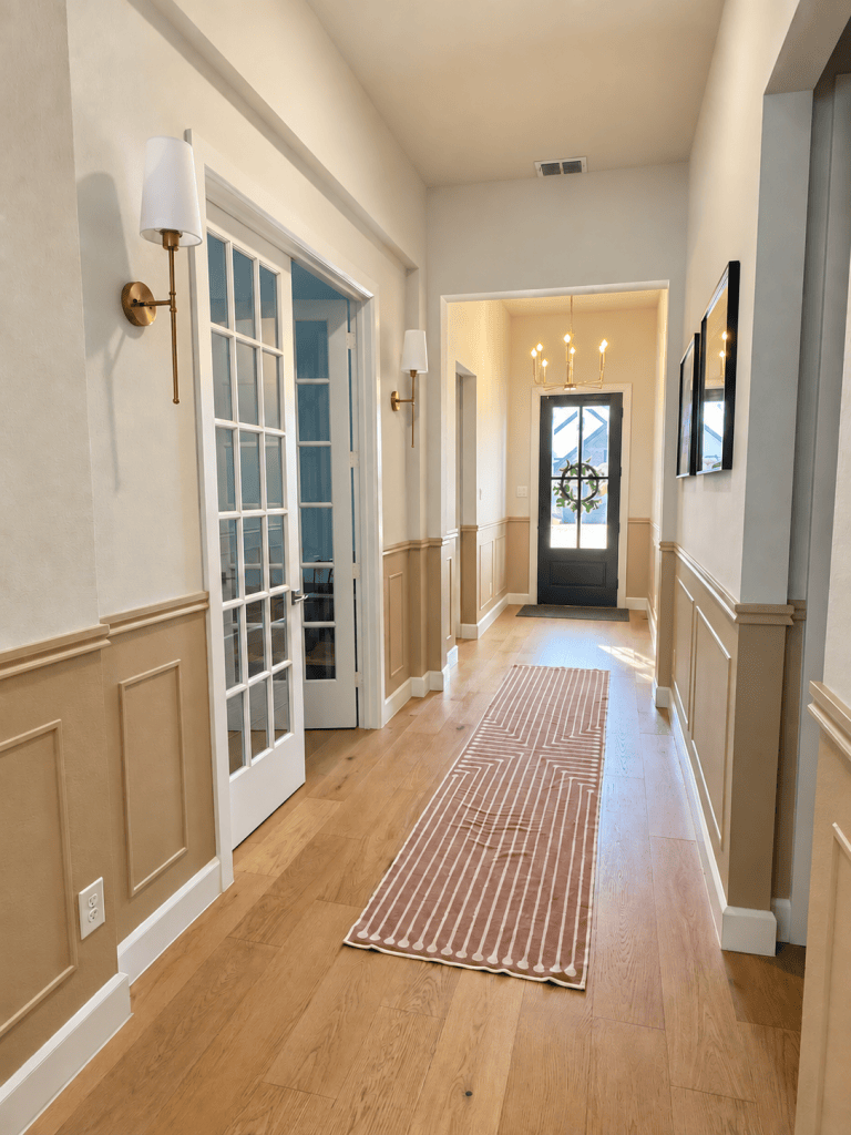 After: long entryway with picture frame wainscoting painted Behr Mushroom Bisque, brass wall sconces, French doors and a black front door