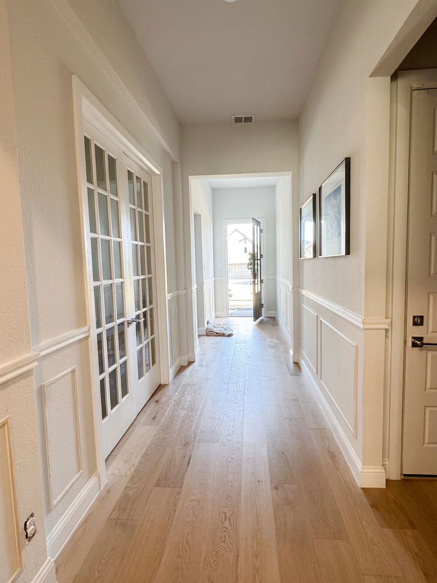 kc-compton-living-entryway-wainscoting-prep A spacious hallway with light-colored wooden flooring, white walls, and decorative molding. French doors are visible on the left, and the hallway leads to an open doorway at the end, with natural light coming through.