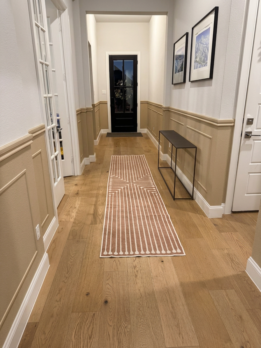 kc-compton-living-entryway-wainscoting-painted-mushroom-bisque A well-lit hallway featuring wooden flooring, light-colored walls, and a decorative area rug with a geometric pattern. There is a black front door at the end, with framed pictures on the walls and a slim console table on one side.